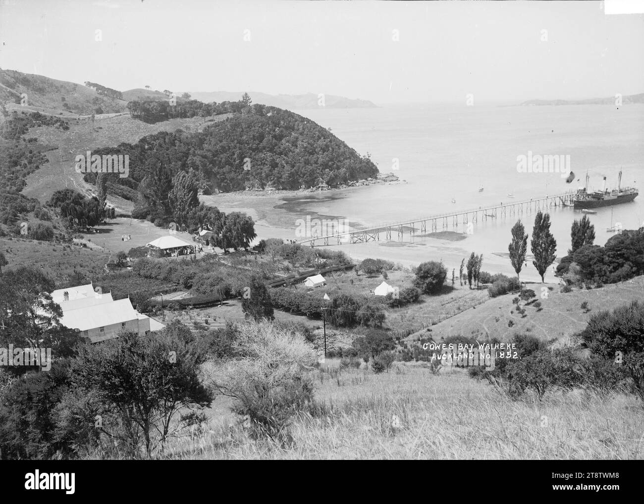 Ferry at the wharf, Cowes Bay, Waiheke Island, View of Cowes Bay taken from a high vantage point above the bay and looking north. Pakatoa Island is just visible in the far distance on the right. A ferry is moored at the wharf. The Cowes Bay boarding house / hotel? is in the immediate foreground on the left. People can be seen crowded around the pavilion or passenger waiting room (?) in the middle distance near the end of the wharf, in early 1900s Stock Photo