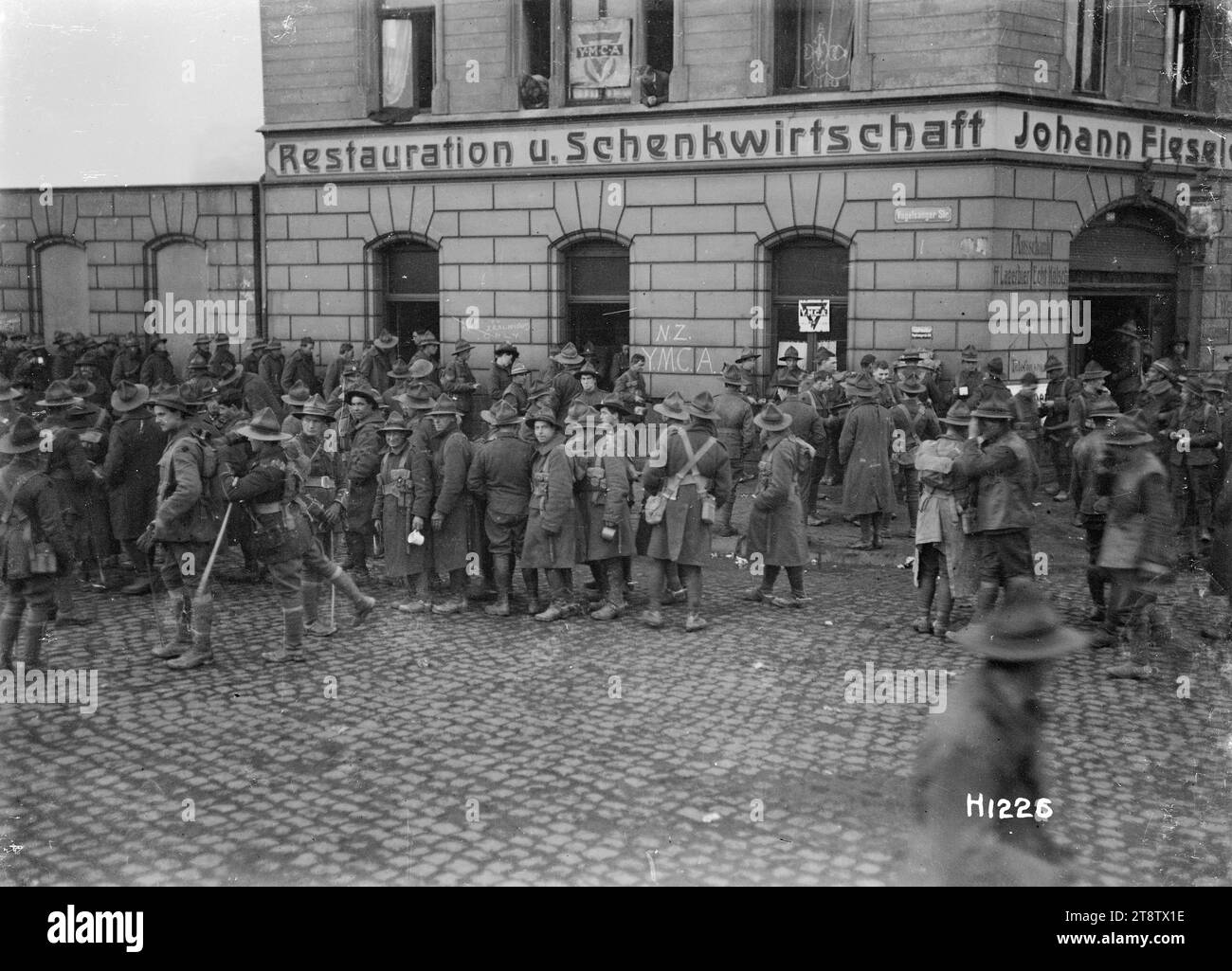 World War I New Zealand troops outside the YMCA in Ehrenfeld, Cologne, A large group of New Zealand soldiers shown mingling on the cobblestones outside the NZ YMCA in Ehrenfeld, a suburb of Cologne in Germany. YMCA notices appear on the window of a building with the German words 'Restauration u. Schenkwirtschaft Johann Fiessler?' on it. 'NZ YMCA' is also written on the wall. Photograph taken after the end of World War I, probably December 1918 Stock Photo