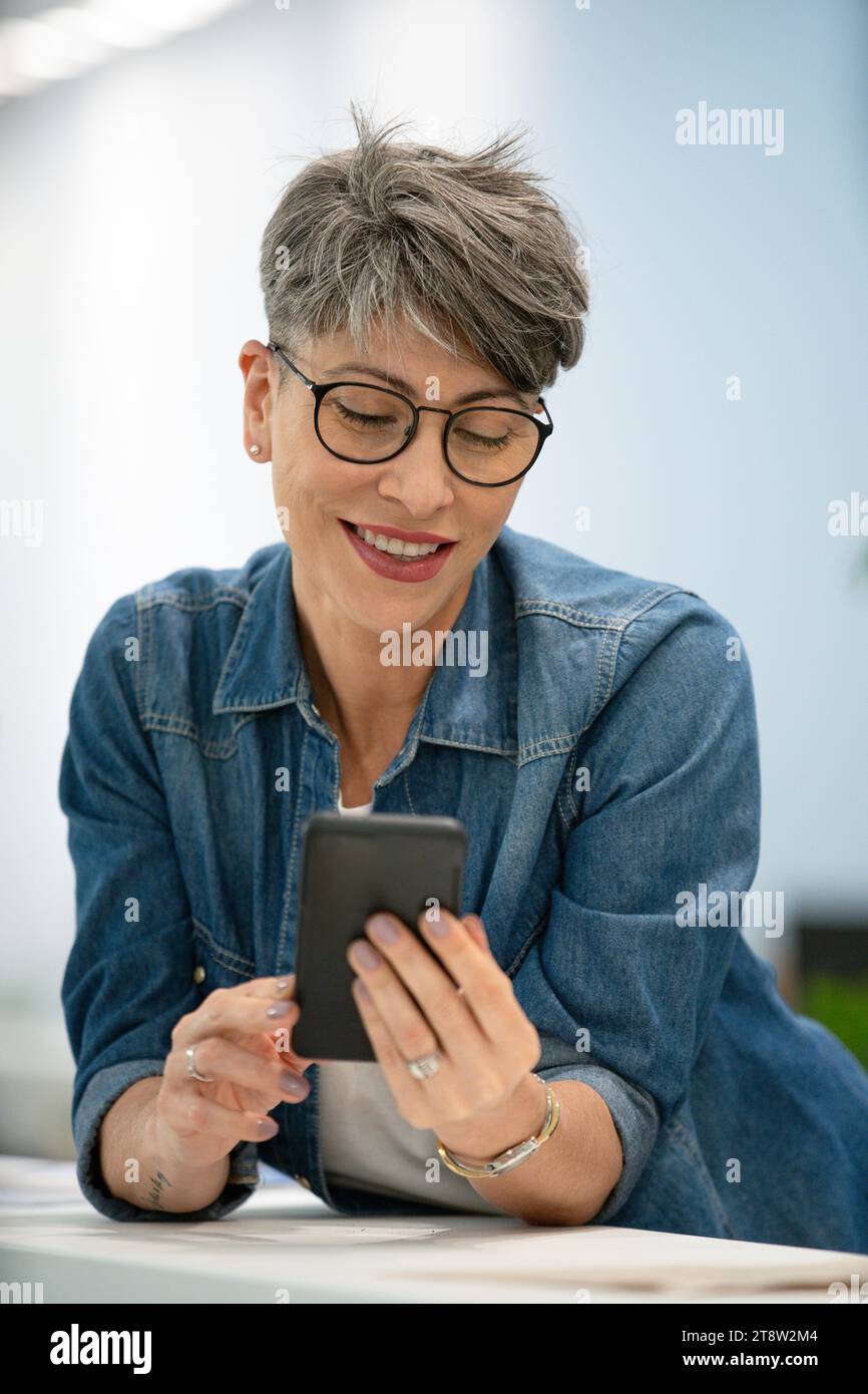 Female entrepreneur scrolling on smart phone while leaning on table Stock Photo