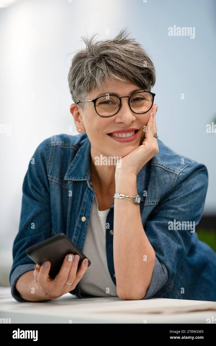 Female entrepreneur holding smart phone while leaning on table looking at the camera Stock Photo