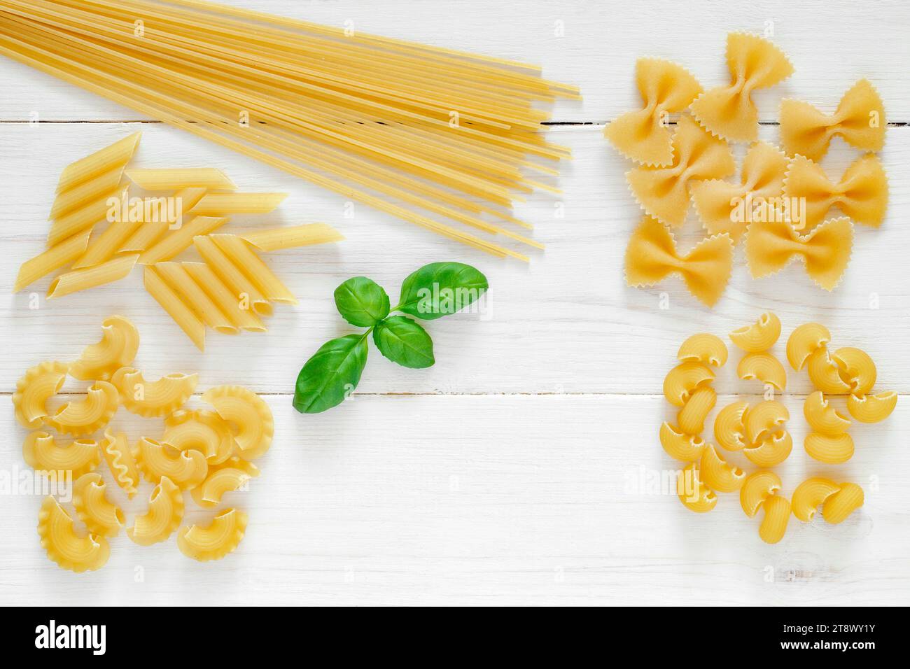 Maccheroni raw, spaghetti, farfalle, pipe rigatoni, penne rigate, cresta di gallo, leaf of basil, on white wooden board background, top view, space to Stock Photo