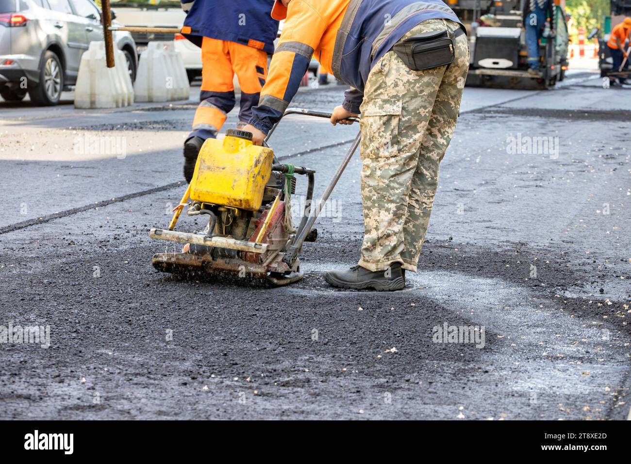 A road service worker compacts fresh asphalt on a section of road with a worn-out gasoline vibrating plate around a sewer manhole. Copy space. Stock Photo