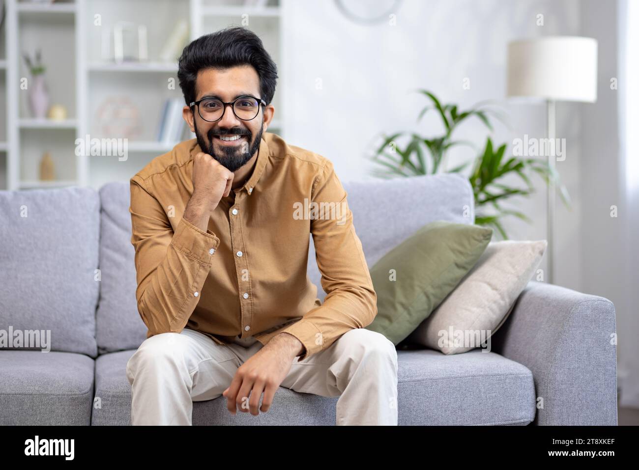 Portrait of a young Indian man sitting on the sofa at home, resting his hand on his chin, confidently and smilingly looking at the camera. Stock Photo