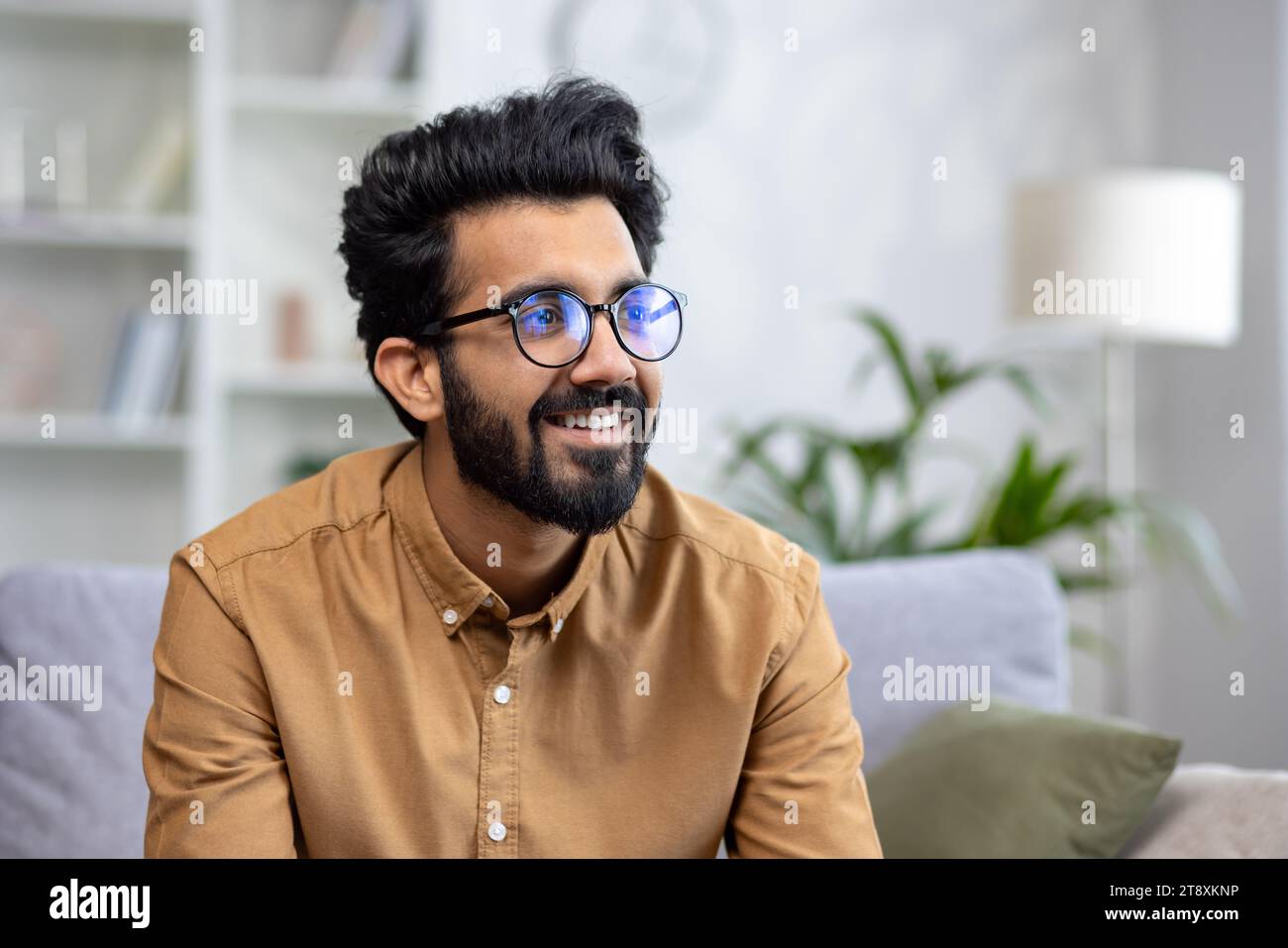 Close-up photo of a young Indian man in glasses sitting on the couch at home and looking to the side with a smile. Stock Photo