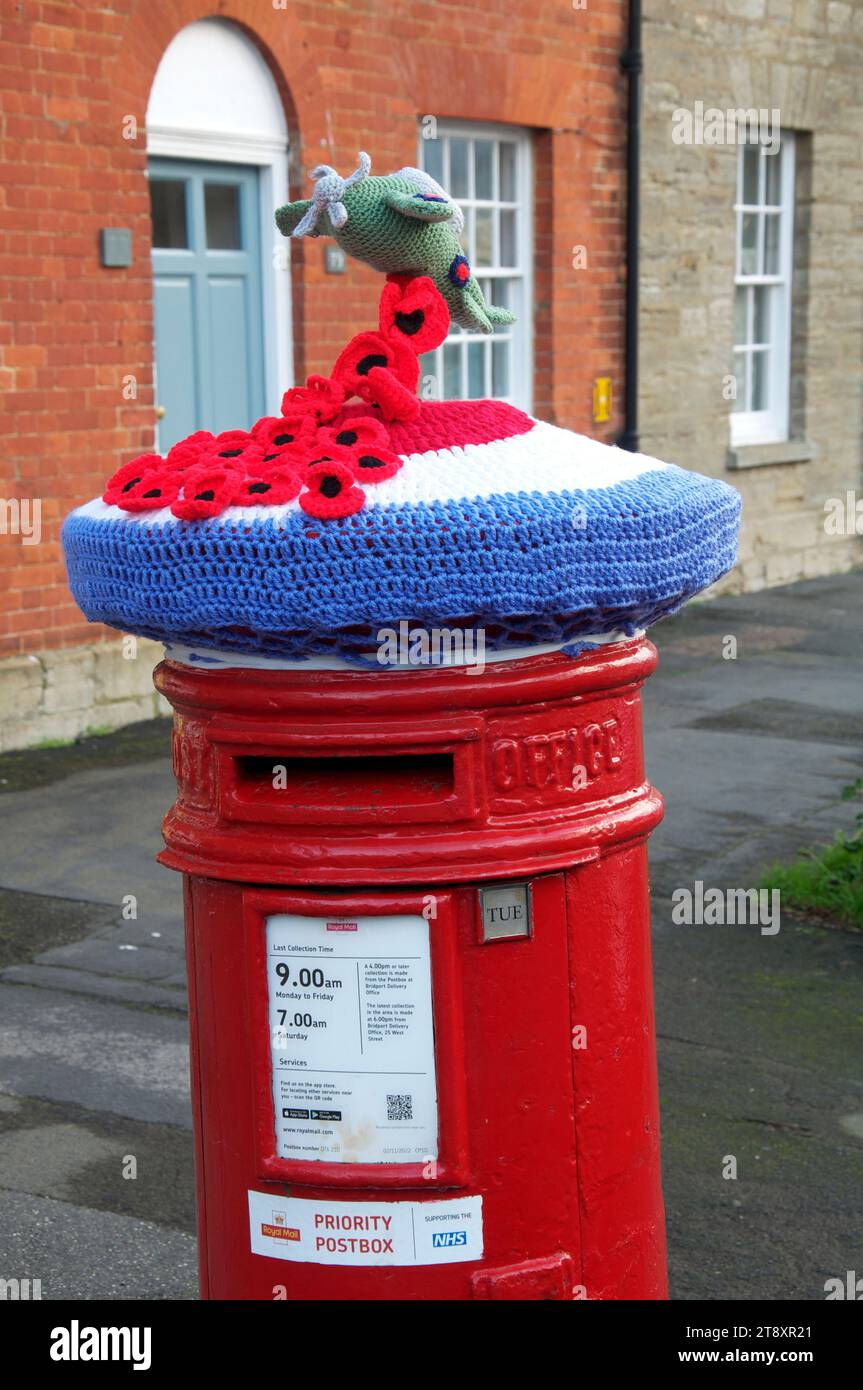 A knitted woollen hat depicting an RAF roundel supporting a crocheted Spitfire and a cascade of poppies, covers an old red pillar box. Remembrance Day. Stock Photo