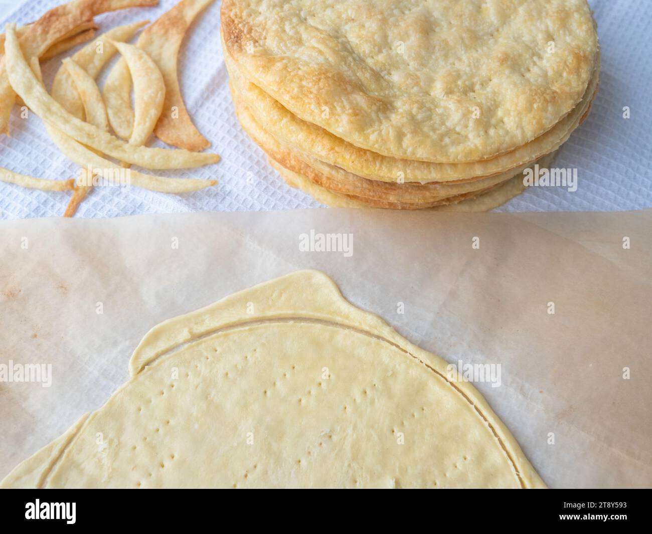 Cooking a shortcrust puff pastry Napoleon cake with a pile of baked golden thin buttery layers, dough scraps, and raw layer rolled out on baking paper Stock Photo
