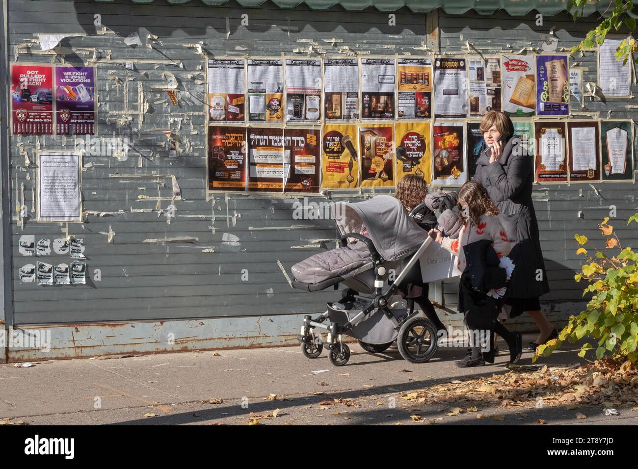 An orthodox Jewish woman and her 3 children walk on a side street past community billboards & notices in Williamsburg, Brooklyn, New York. Stock Photo