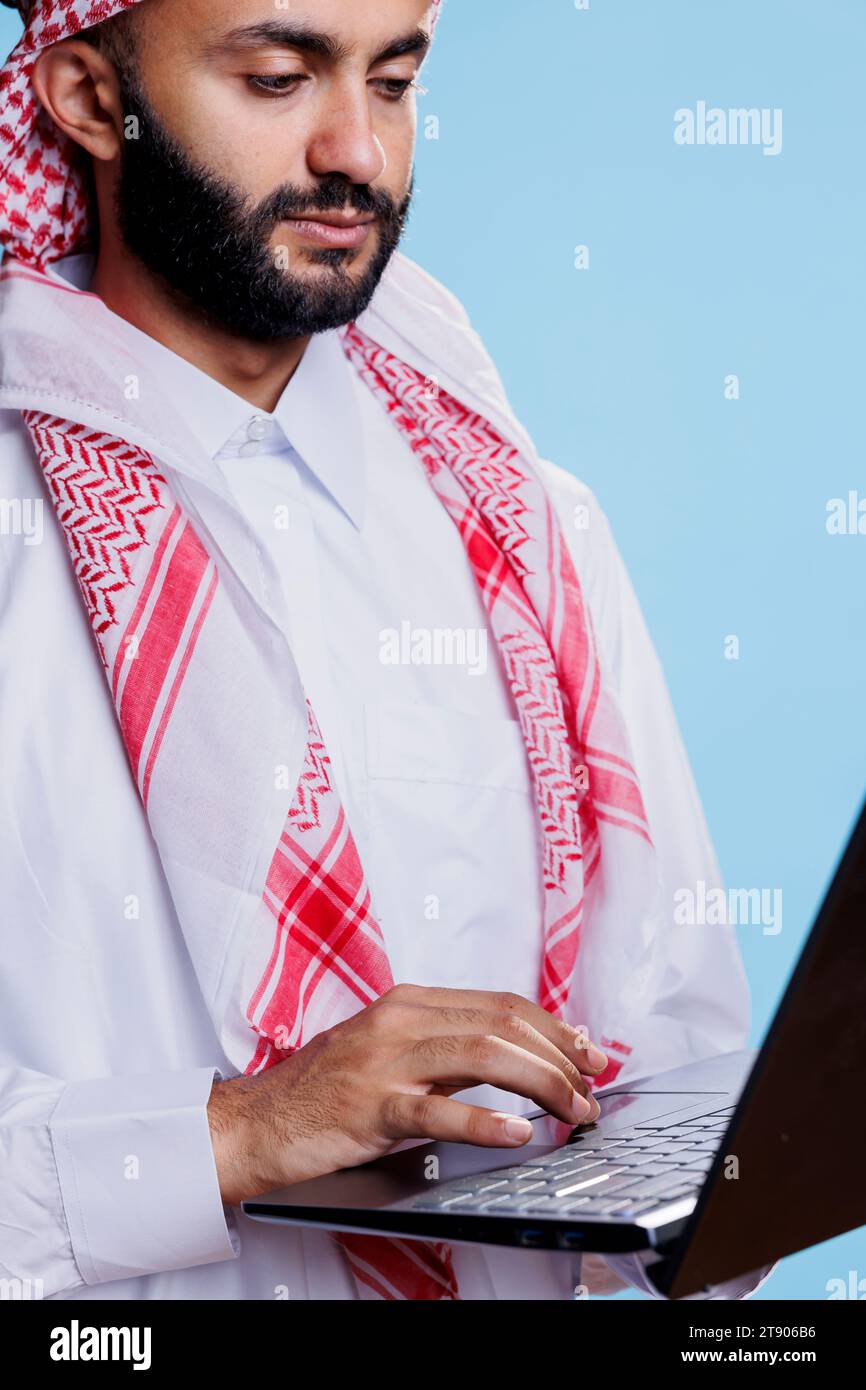 Man dressed in traditional muslim thobe and using laptop touchpad closeup. Arab person wearing islamic attire, holding portable computer and scrolling page while browsing internet Stock Photo