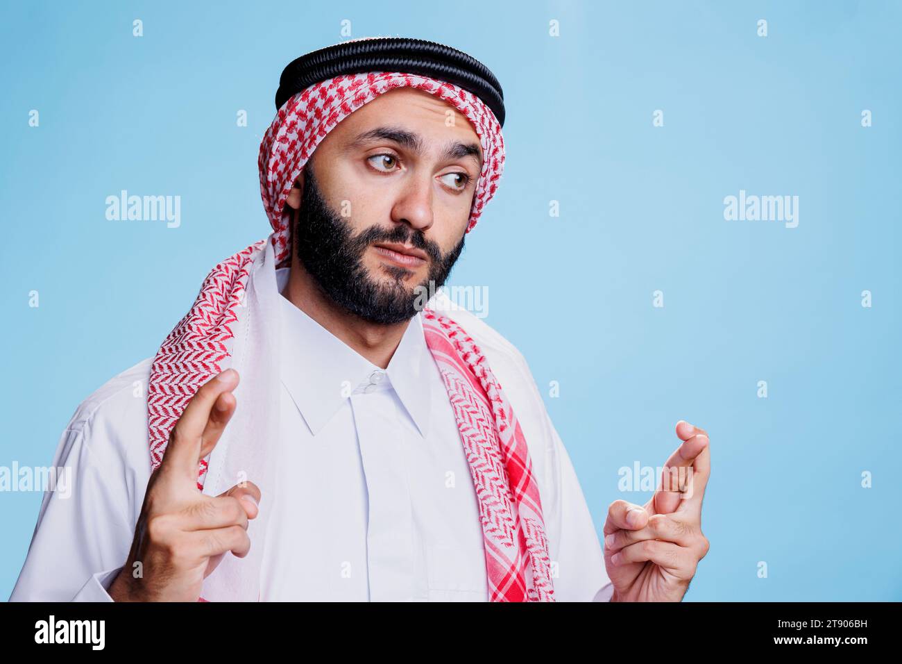 Arab man wearing traditional thobe and headscarf crossing fingers for good luck. Person dressed in islamic cultural clothes showing superstitious gesture, pleading for success Stock Photo