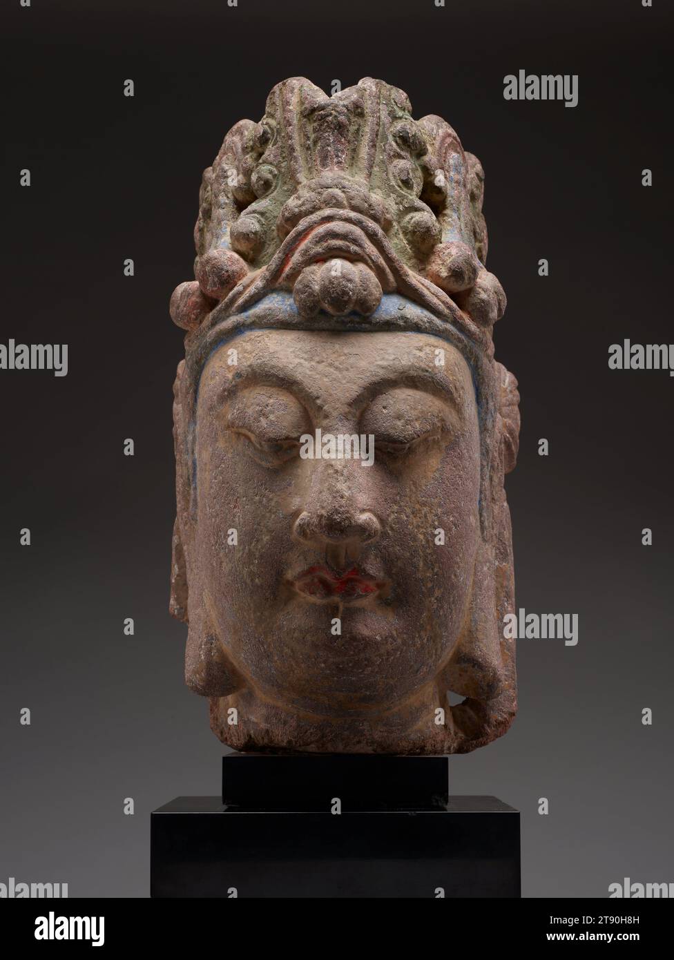 Head of a Bodhisattva, late 6th century, 16 1/4 × 9 7/16 × 11 5/8 in. (41.28 × 23.97 × 29.53 cm) (without attached base), Sandstone, pigments, China, 6th century, This sculpture was originally the head of a standing figure of a bodhisattva—a being who has postponed its own passage to nirvana in order to guide others to salvation. Carved fully in the round, with a serene expression and gracefully curved downcast eyes, it wears an elaborate crown that is secured by knotted ribbons that hang down behind its ears. These characteristic knots of scarves help date the sculpture to the Sui dynasty Stock Photo