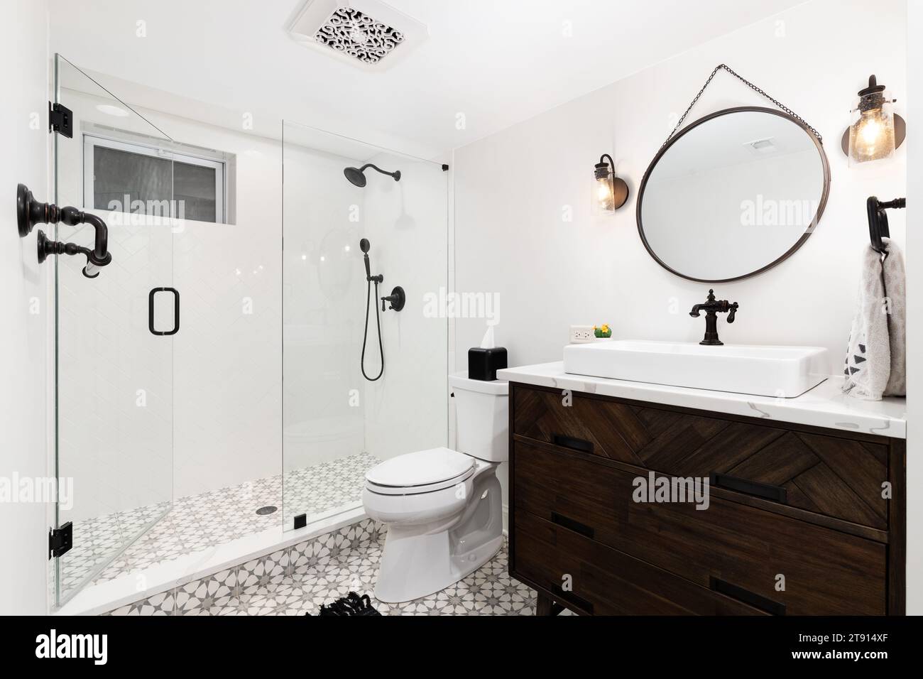A cozy bathroom with a dark wood cabinet and vessel sink, an mosaic pattern tile flooring, and herringbone shower tile walls. Stock Photo