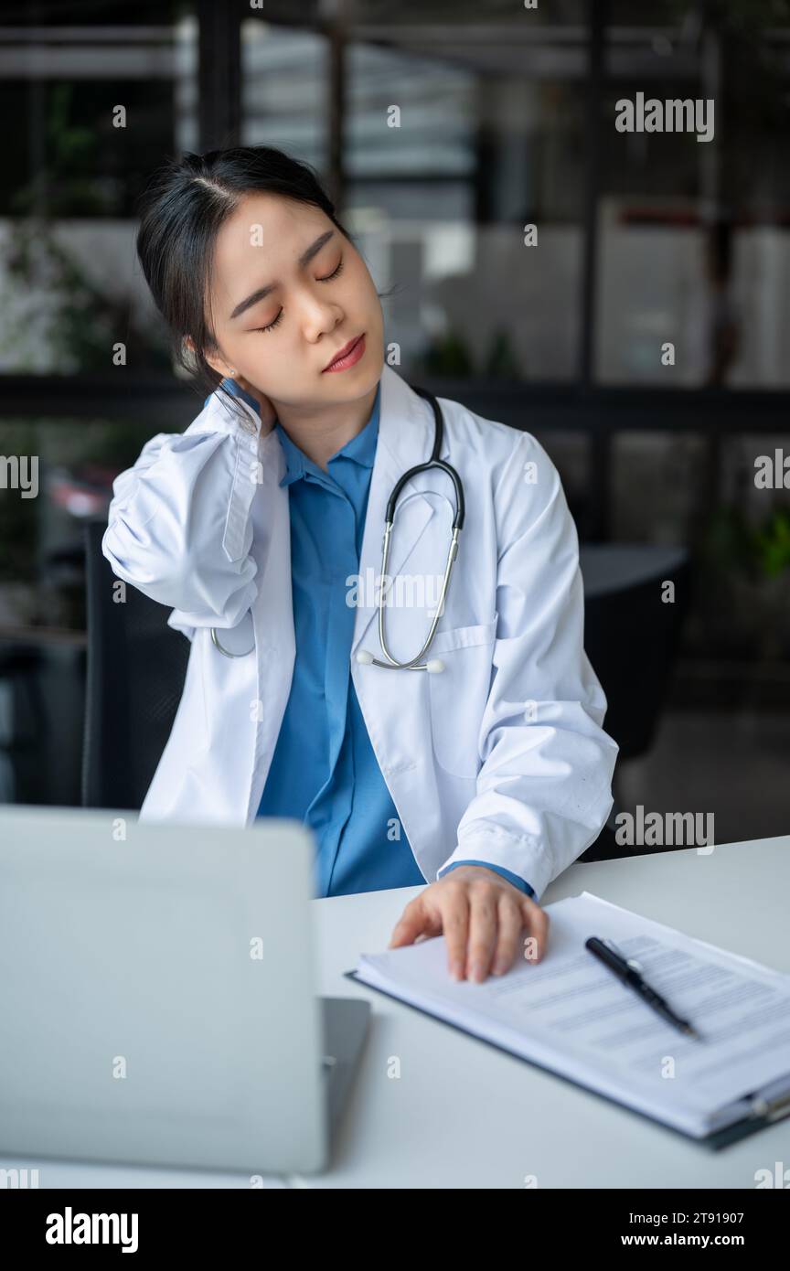 A tired and overworked young Asian female doctor is massaging her neck ...