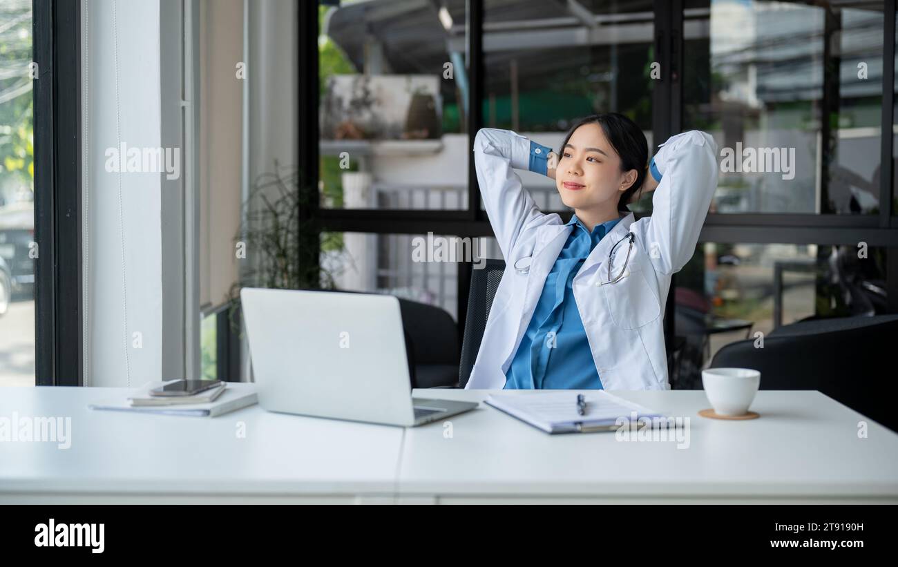 A relaxed and carefree Asian female doctor is putting her hands behind her head, looking outside, daydreaming and feeling fresh while taking a break a Stock Photo