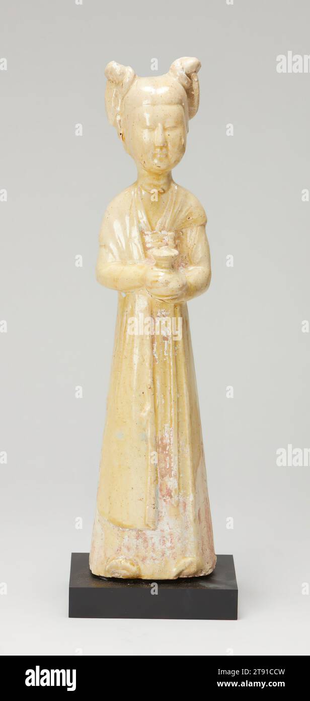 Female Attendant, one of five, 7th century, 12-3/16 x 3 x 2-7/8 in. (31.0 x 7.6 x 7.3 cm), Earthenware with straw-colored glaze and painted pigments, China, 7th century, Shown standing in a quiet and respectful attitude holding small, pear-shaped covered vases, these figures of young women represent court attendants. Dressed in similar attire, with aquamarine blue scarves, red-striped skirts, and high-waisted dresses, they all have the full, round faces, slender figures, and double topknots that were fashionable during the early Tang dynasty Stock Photo