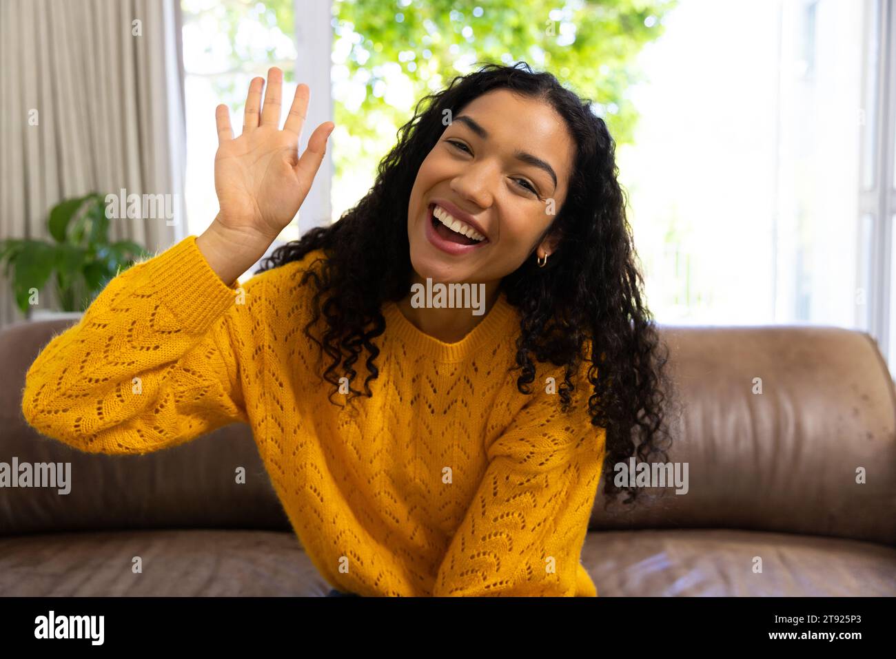 Happy biracial woman in yellow sweater making video call and waving at home Stock Photo