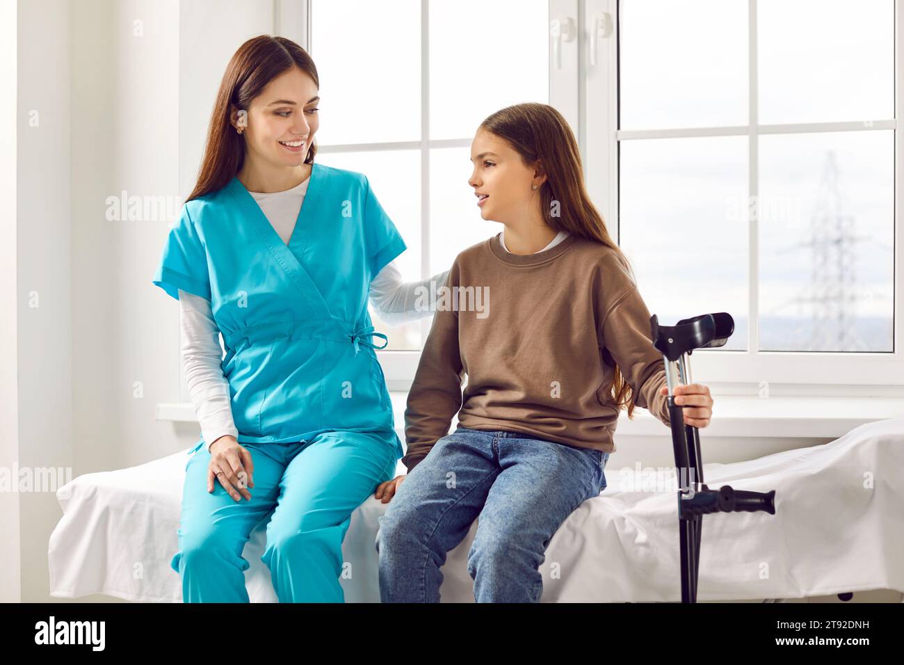 Young friendly nurse talking with a child girl patient sitting on the couch in clinic with crutches Stock Photo