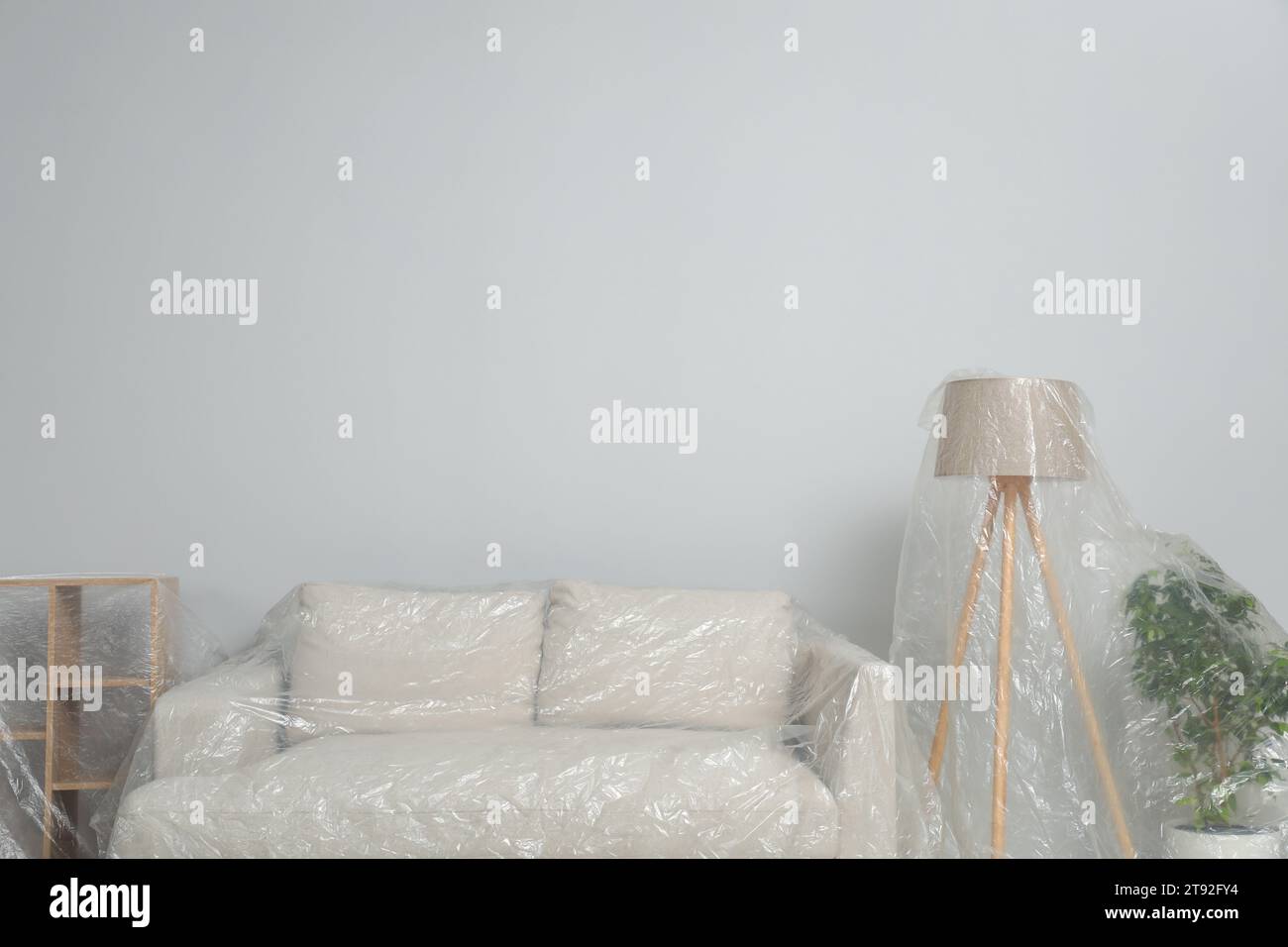 Sofa, shelving unit, lamp and houseplant covered with plastic film near light wall indoors. Space for text Stock Photo