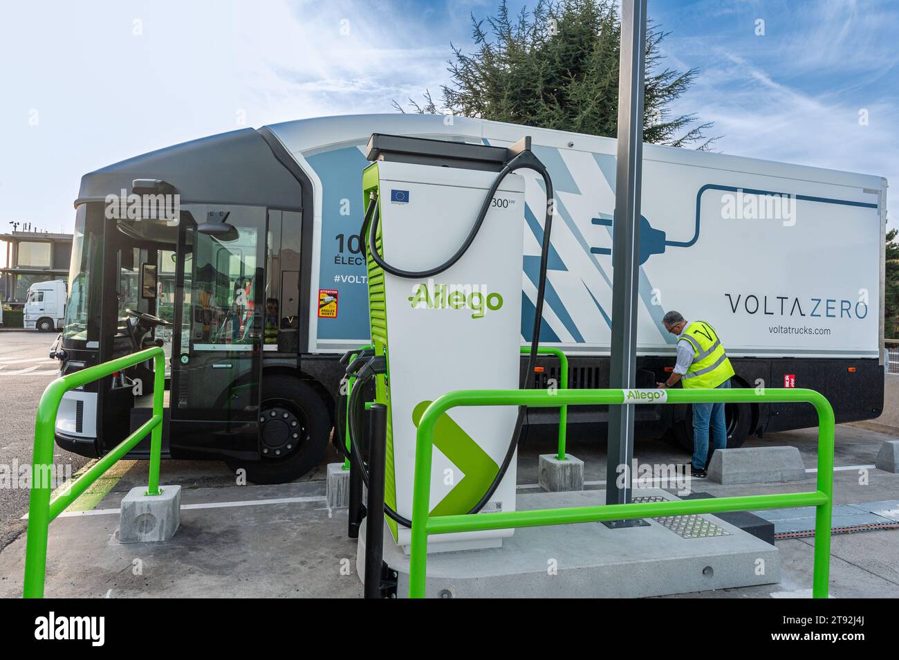 A Volta Zero truck is parked at an Allego charging station. A man in reflective jacket inspects it with a tree and clear blue sky in the background Stock Photo