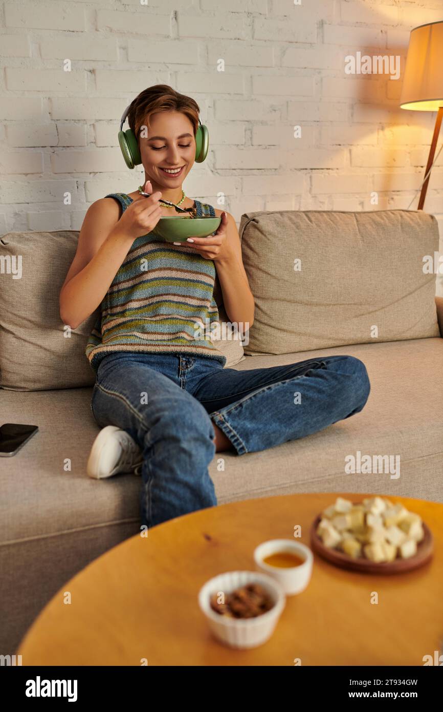 joyful woman in headphones having vegetarian dinner near on couch near table with plant-based meal Stock Photo