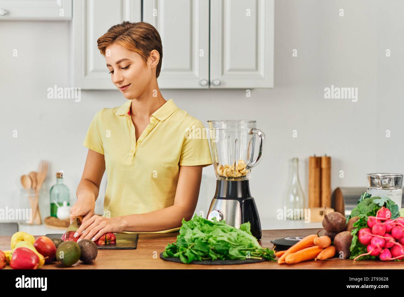 young vegetarian woman cutting apple near electric blender and assortment of vegetables and fruits Stock Photo