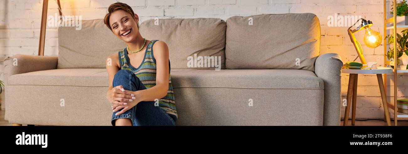 joyous young woman sitting near couch in cozy living room and looking at camera, horizontal banner Stock Photo