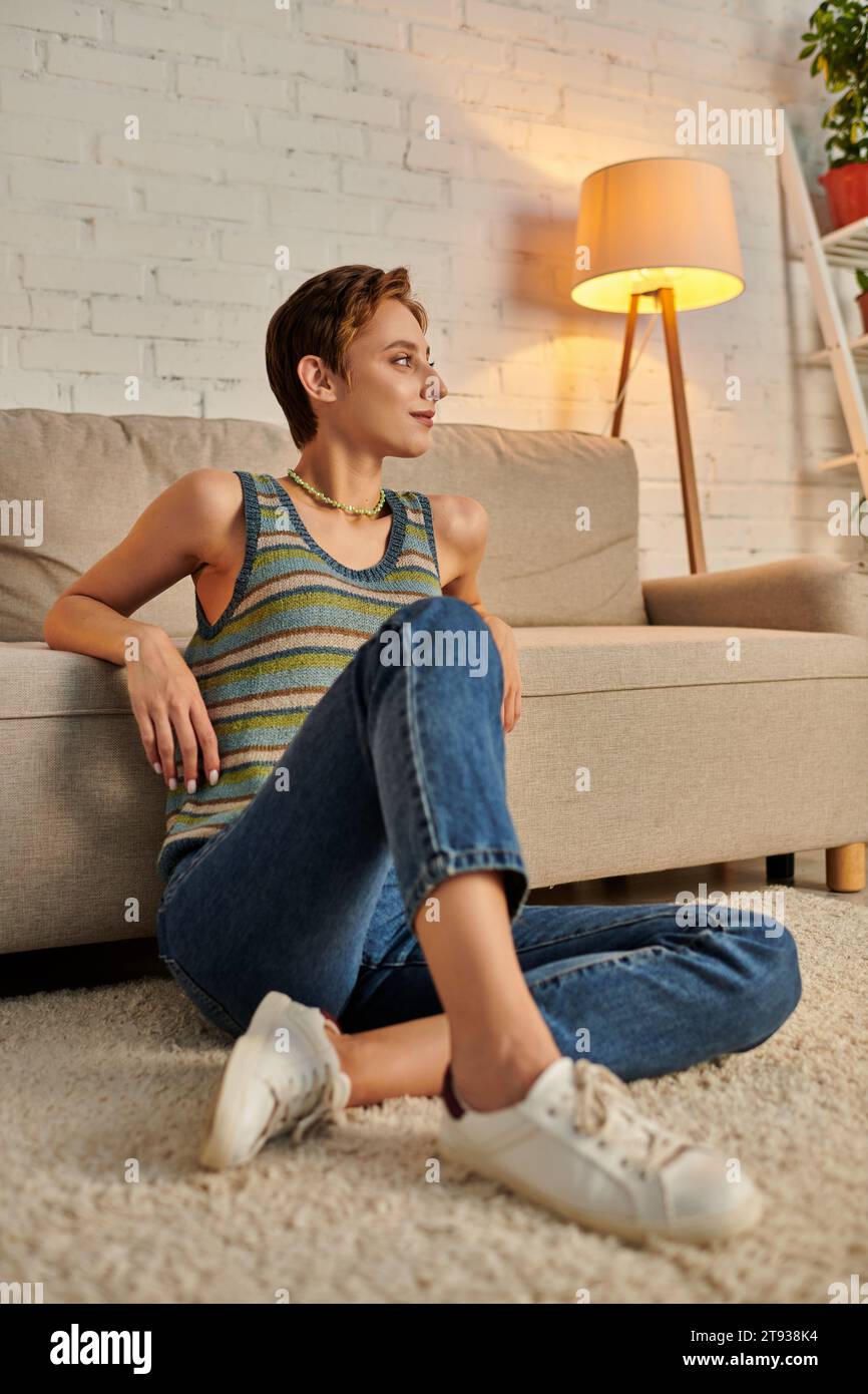positive and dreamy young woman looking away while sitting on floor near couch in living room Stock Photo