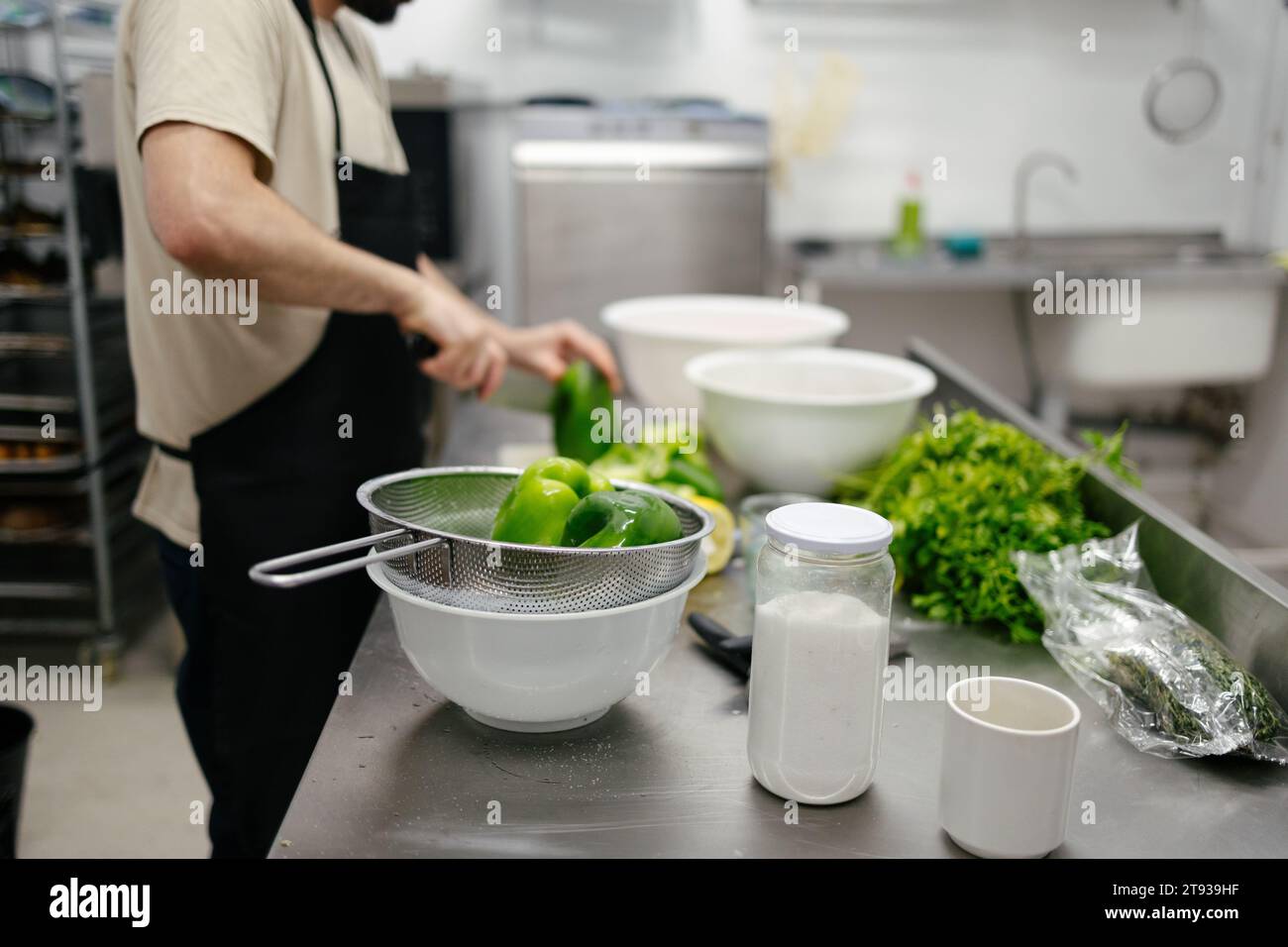 Close-up of green peppers while in the background there is an out-of-focus and unrecognizable chef cutting peppers in a professional kitchen Stock Photo