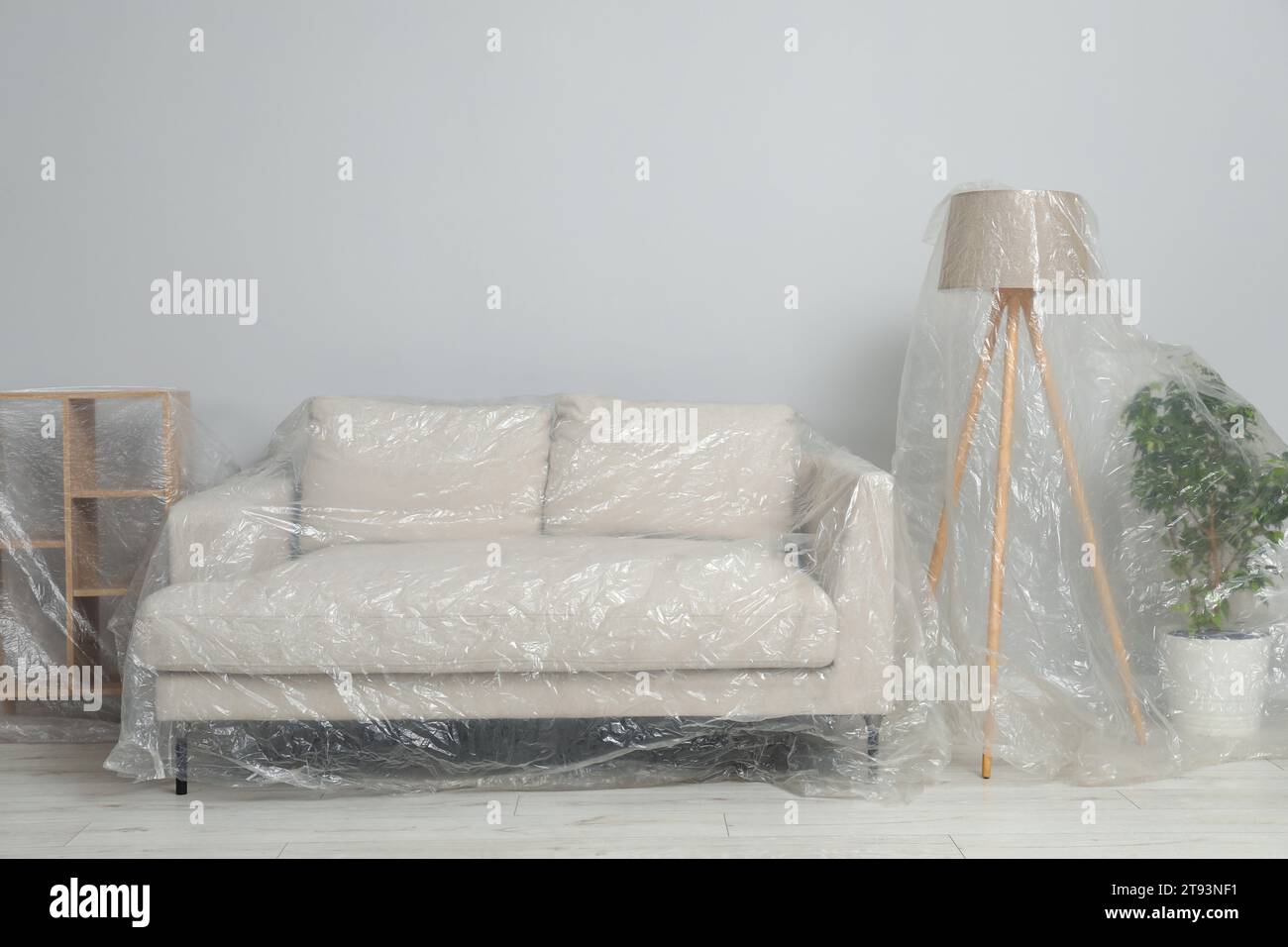 Sofa, shelving unit, lamp and houseplant covered with plastic film near light grey wall indoors Stock Photo