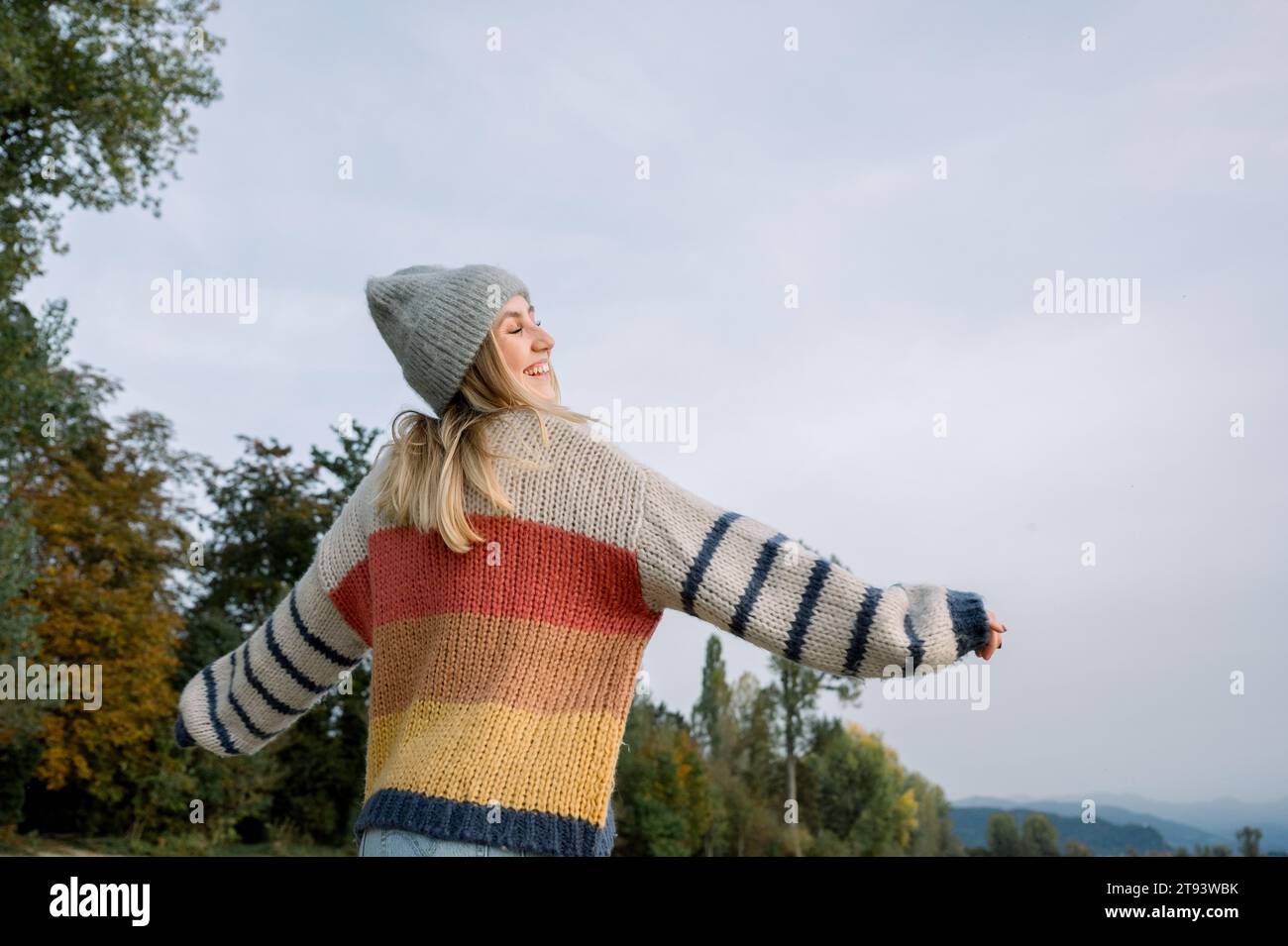 From below of young female in warm sweater and knitted hat standing against green trees and cloud while enjoying sunny day in countryside on winter ti Stock Photo