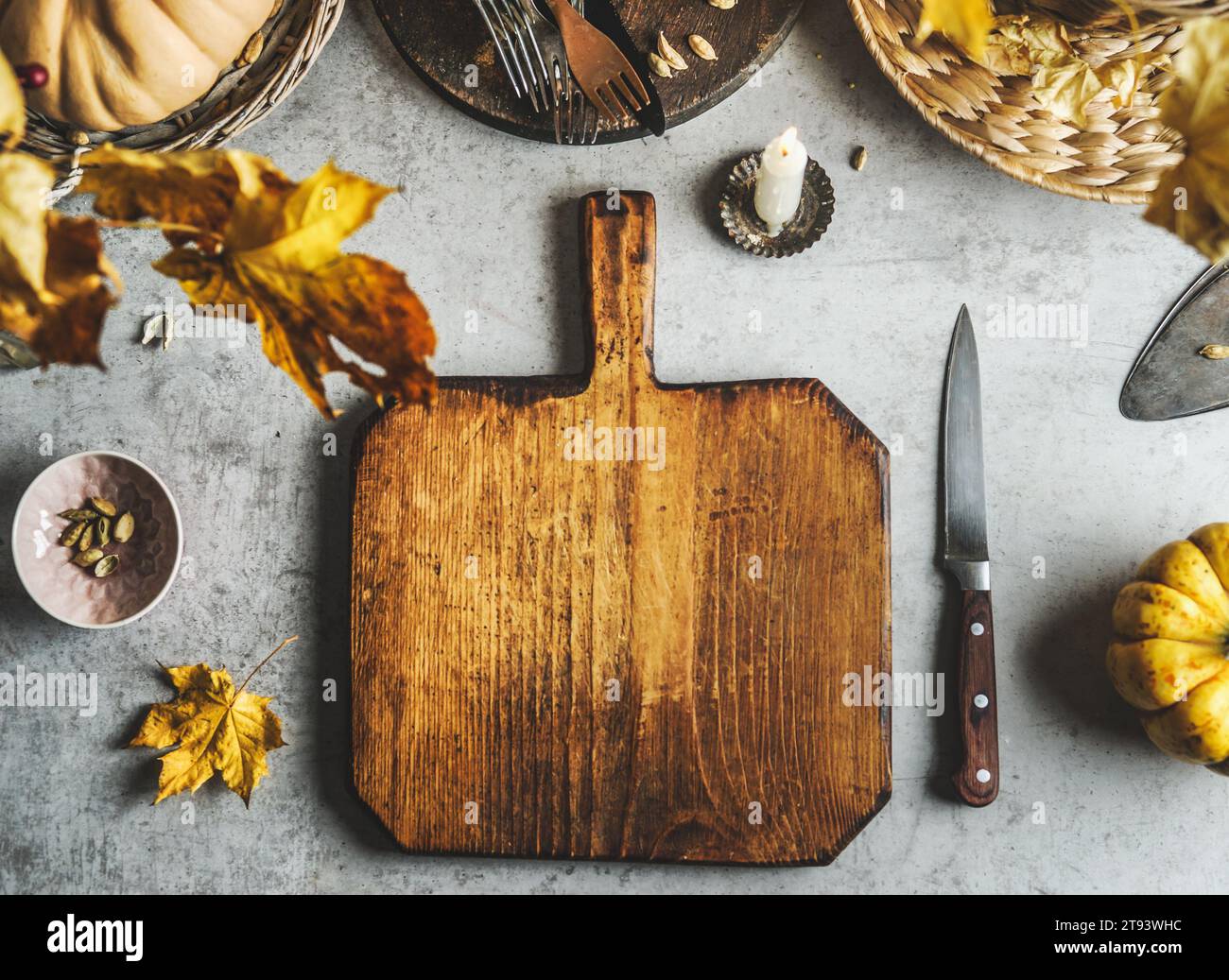 Food background with empty wooden cutting board, pumpkin, knife, autumn leaves and utensils at grey table. Cooking at home with seasonal ingredients. Stock Photo