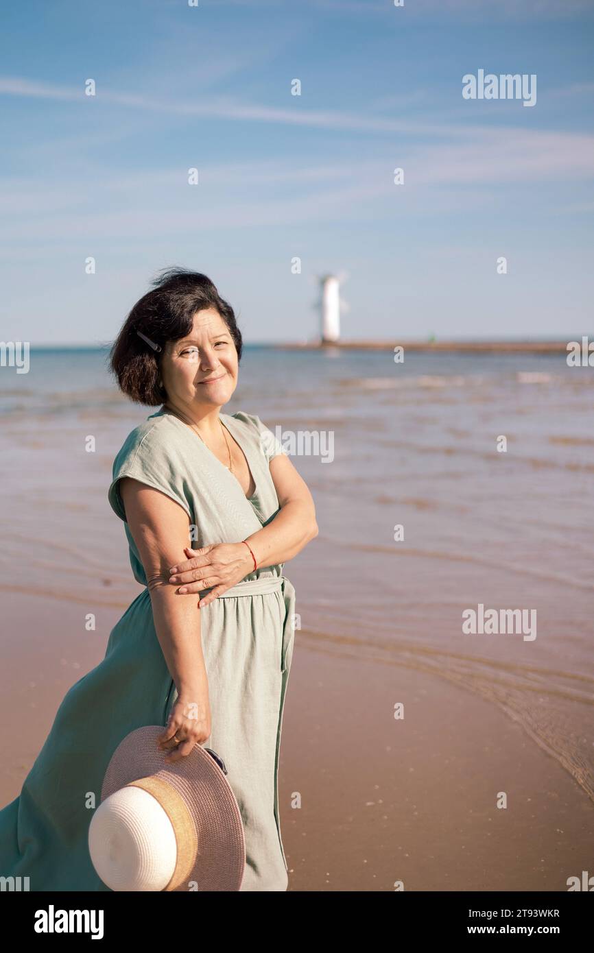 A woman in a flowing dress stands on a sandy beach, from the sun with a ...