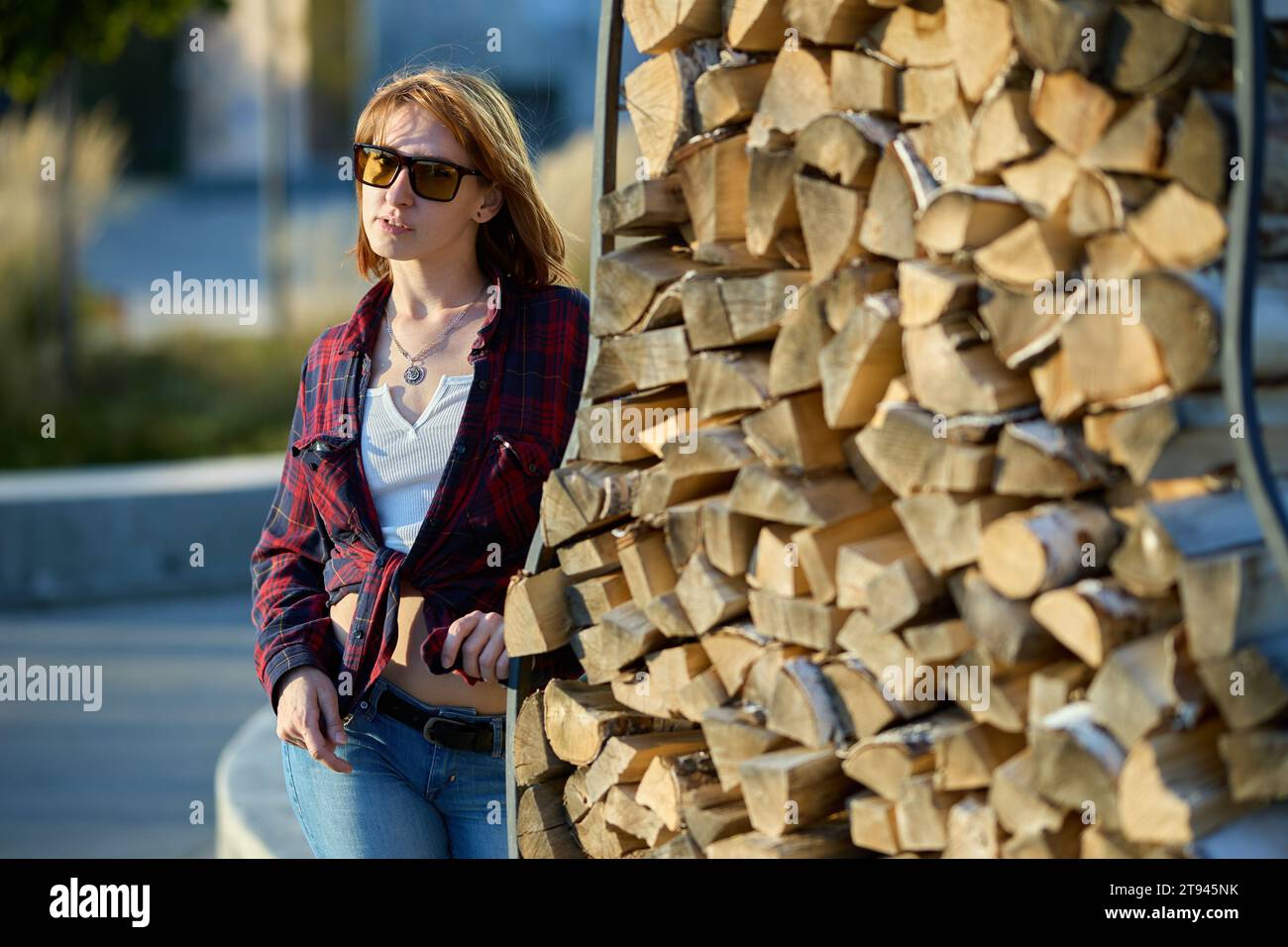 A fashionable redhead woman enjoying an autumn evening in the city. She's wearing a trendy red jacket, a white t-shirt, and denim jeans, making a bold Stock Photo