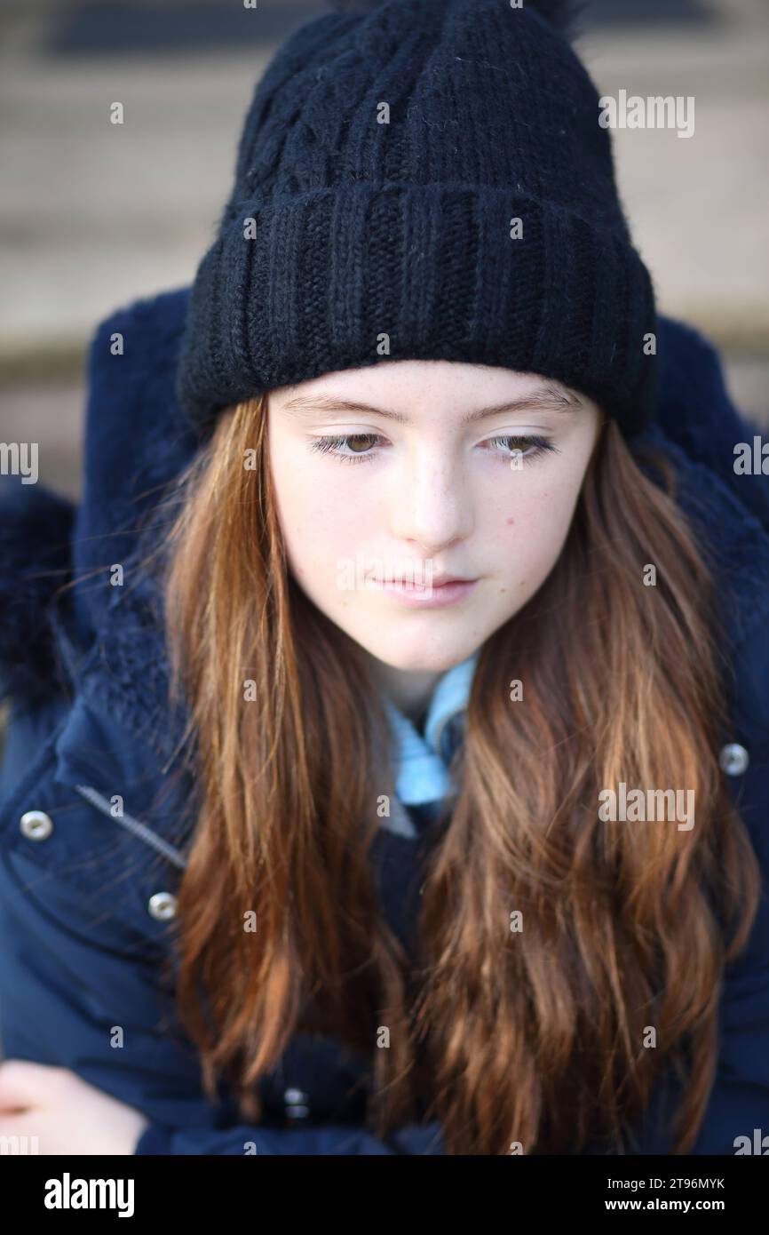 Girl sitting on step wearing woolly hat and coat and scarf trying to keep warm, looking down and upset Stock Photo
