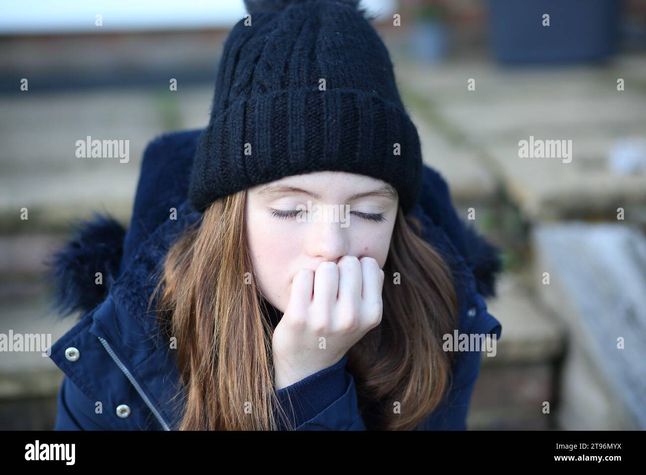 Teen girl sitting on step wearing woolly hat and coat and scarf trying to keep warm with her eyes closed and hand on chin over mouth Stock Photo