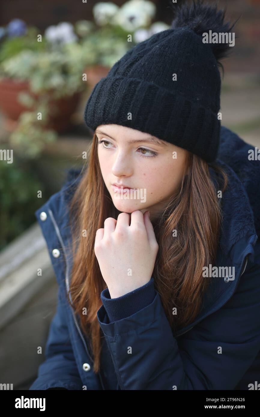 Girl sitting on step wearing woolly hat and coat and scarf trying to keep warm, looking thoughtful with hand under chin Stock Photo
