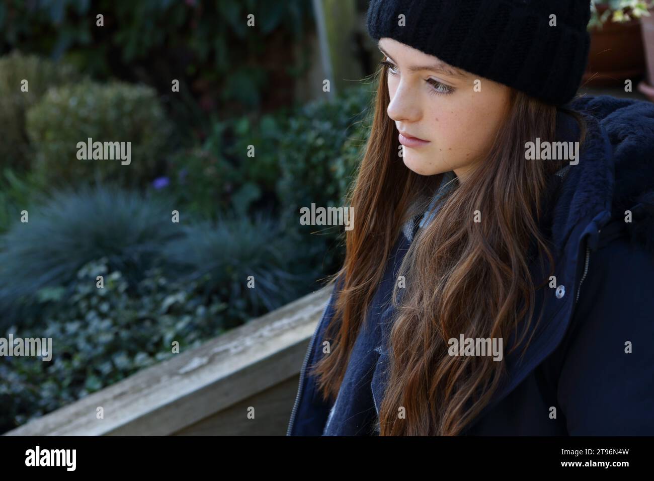 Girl sitting on step wearing woolly hat and coat and scarf trying to keep warm with plants in background Stock Photo