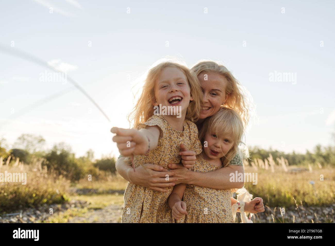 Happy young blond haired woman hugging cute little girls wearing matching dresses and looking at camera against sunlight in countryside Stock Photo