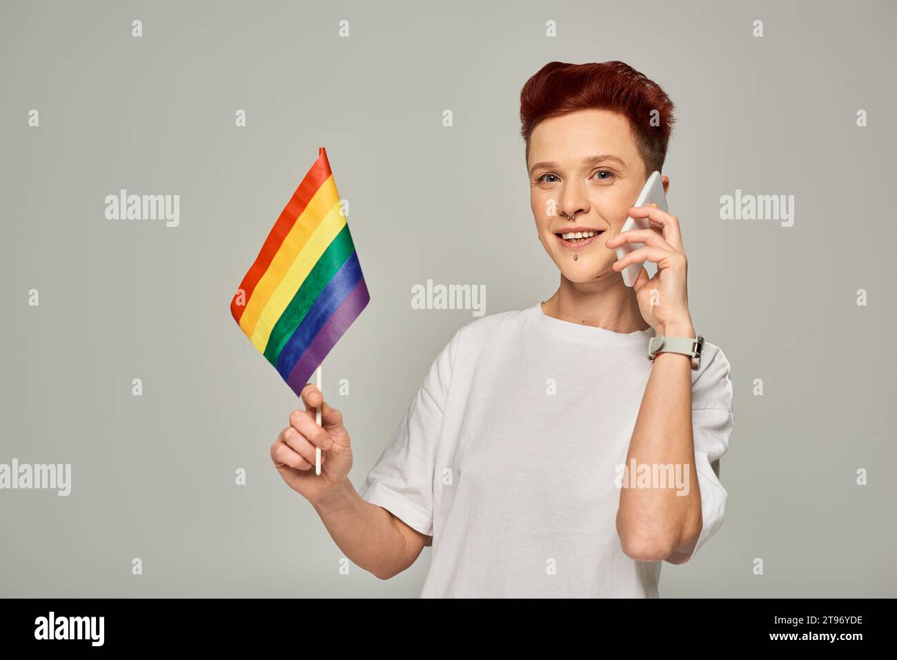 happy redhead non-binary person holding small LGBT flag and talking on mobile phone on grey Stock Photo