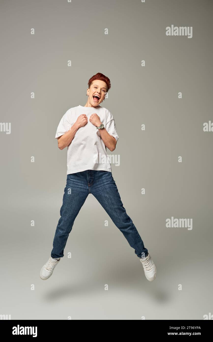 full length of overjoyed non-binary person in white t-shirt and jeans levitating on grey backdrop Stock Photo