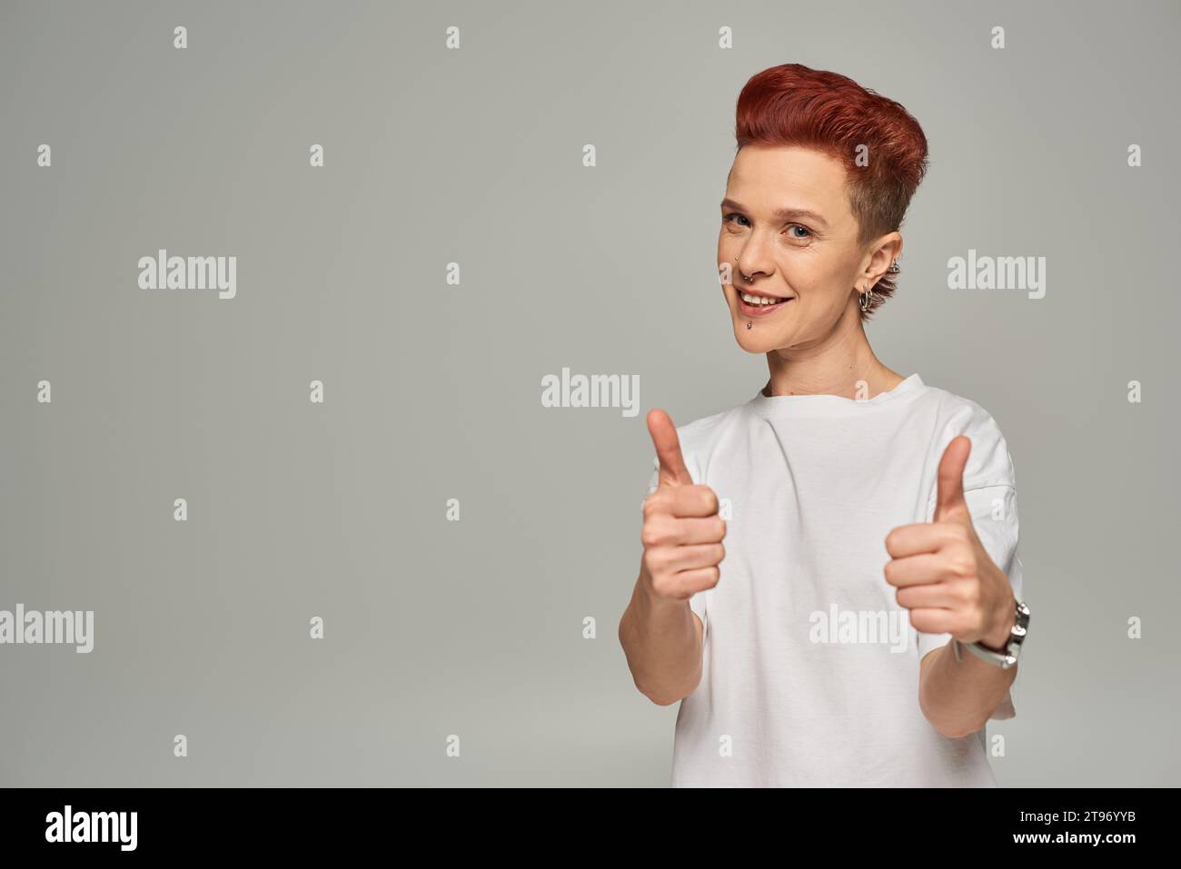 happy redhead non-binary person in white t-shirt showing thumbs and smiling at camera on grey Stock Photo