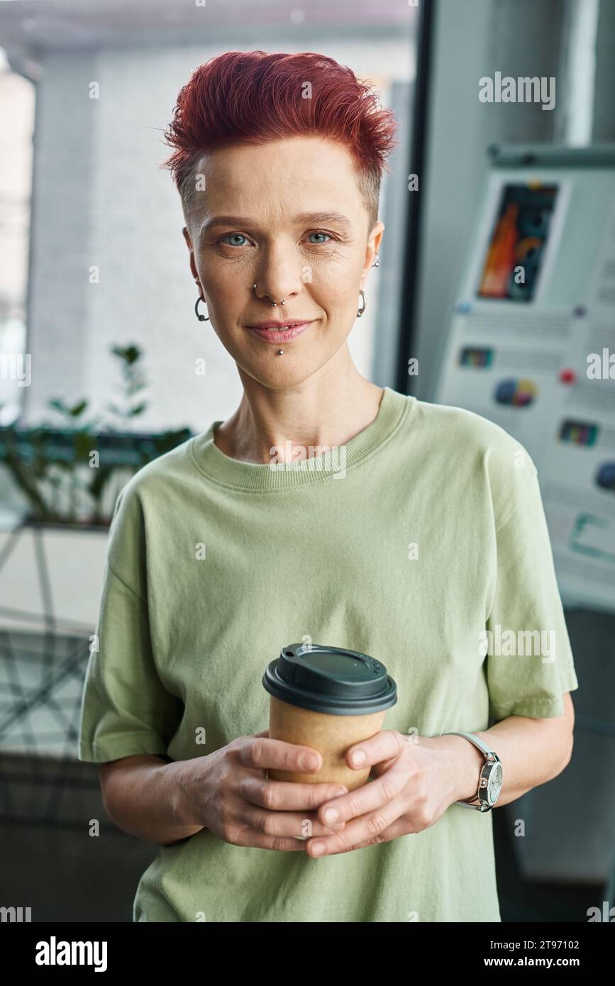 smiling non-binary person with takeaway drink in paper cup looking at camera in modern office Stock Photo