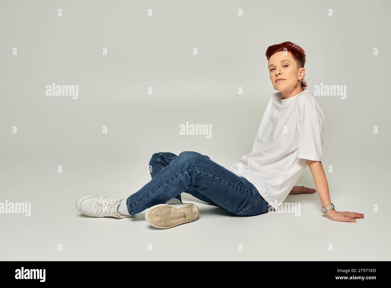 stylish redhead non-binary person in white t-shirt and jeans sitting and looking at camera on grey Stock Photo