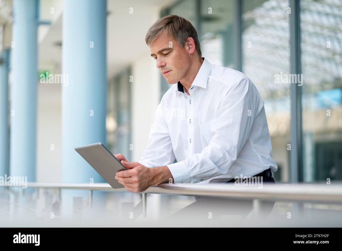 Businessman leaning on railing in office building using digital tablet Stock Photo