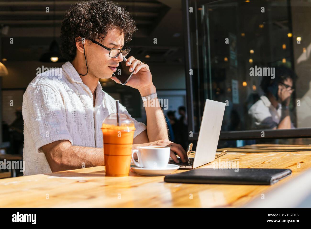 Young freelancer talking on smart phone and using laptop in cafe Stock Photo