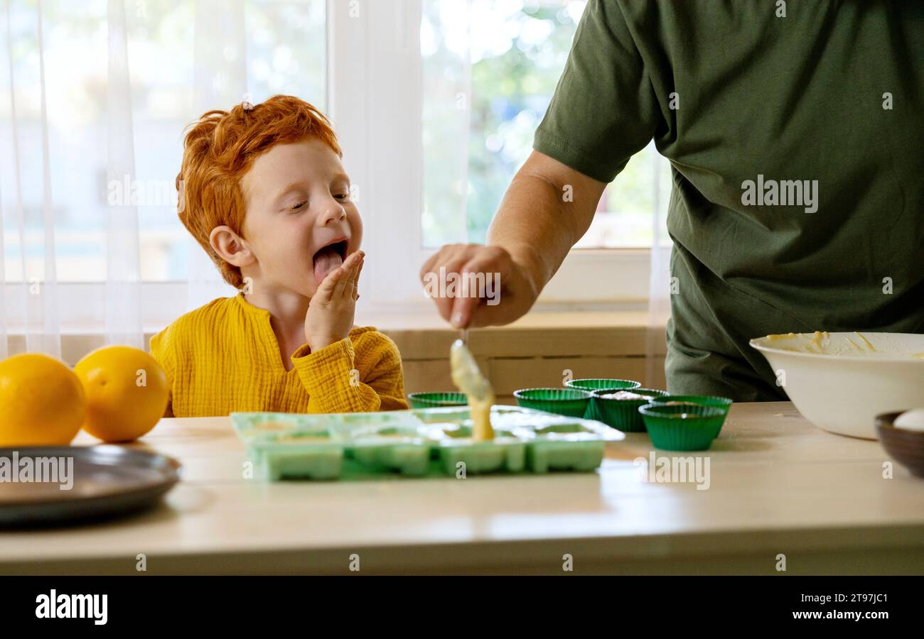 Grandmother putting cupcake batter in molds with grandson licking palm of hand at kitchen Stock Photo