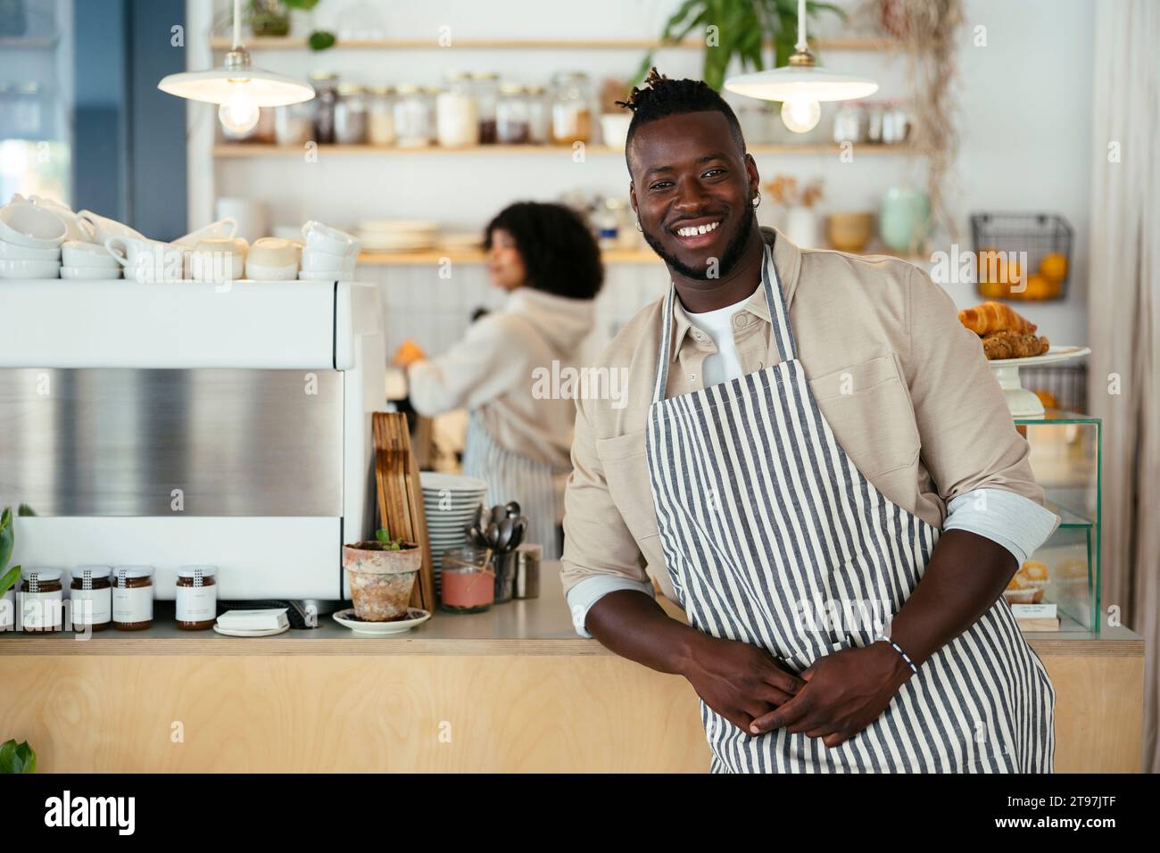 Smiling barista wearing apron standing in front of counter at coffee shop Stock Photo