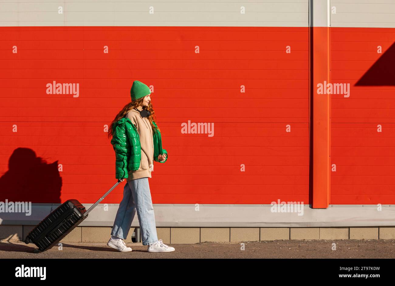 Young woman wearing knit hat walking with wheeled suitcase in front of red wall Stock Photo