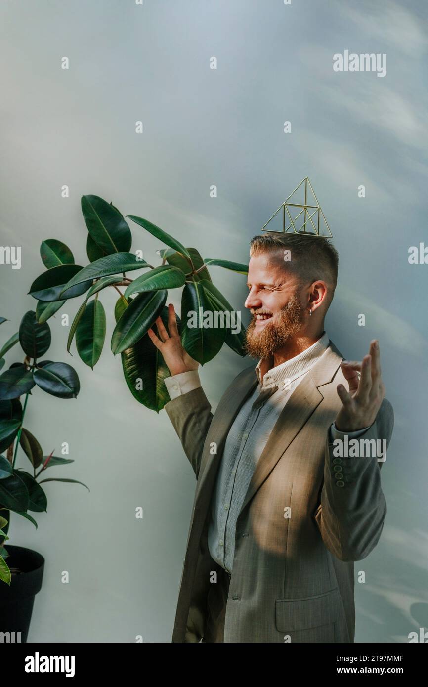 Happy businessman with eyes closed meditating and balancing metal pyramid on head in front of wall Stock Photo