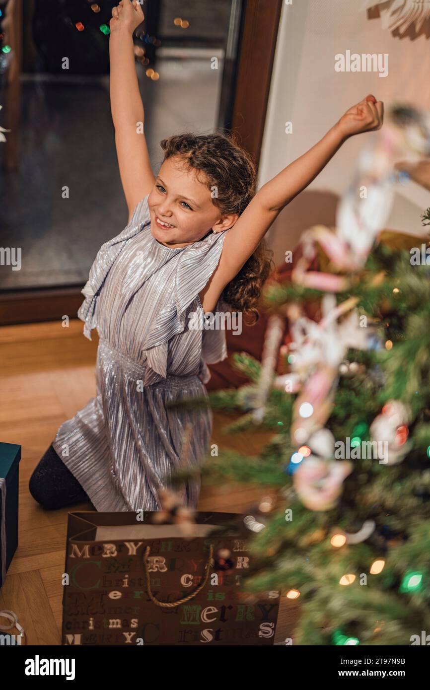 Smiling girl with arms raised kneeling near Christmas tree at home Stock Photo
