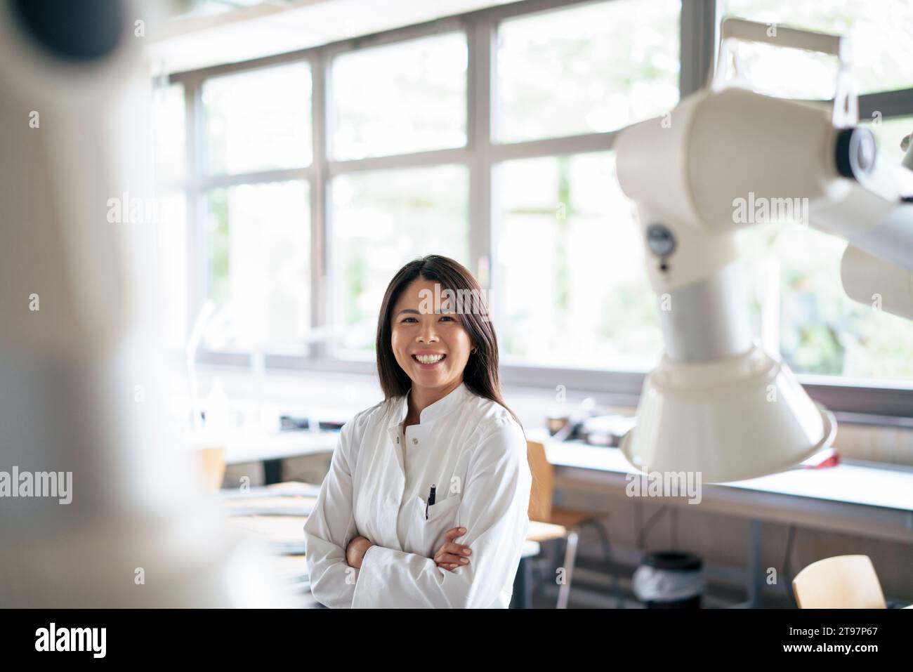 Happy scientist standing with arms crossed near robotic arm Stock Photo